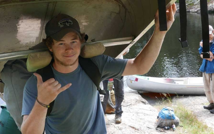 A person balances a canoe on their shoulder and give the camera a "hang loose" sign. 